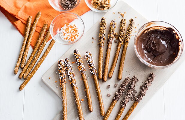 Varillas de pretzel bañadas en chocolate, chispas y dulces, junto a tazones de chocolate y aderezos.