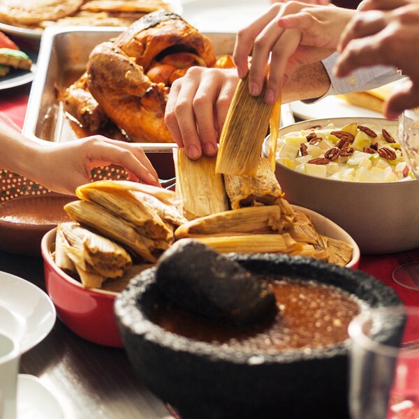 Manos toman tamales de un plato cerca de una bandeja con pavo, un tazón de ensalada de frutas y un mortero lleno de salsa.