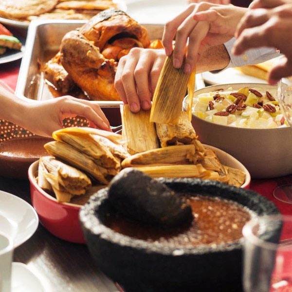 Manos toman tamales de un plato cerca de una bandeja con pavo, un tazón de ensalada de frutas y un mortero lleno de salsa.