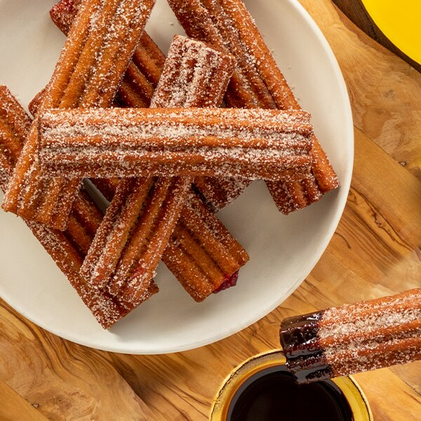Churros azucarados, unos sobre otros, en un plato blanco junto a un churro con chocolate.
