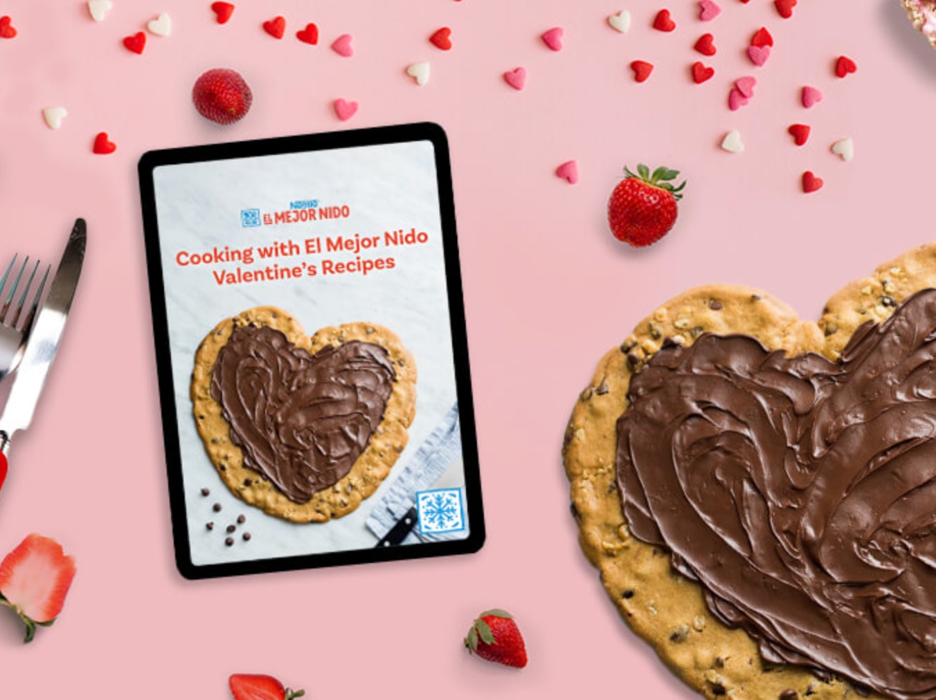 Cookie cake next to tablet with valentines day recipes on pink background with strawberries and hearts and more pink treats.