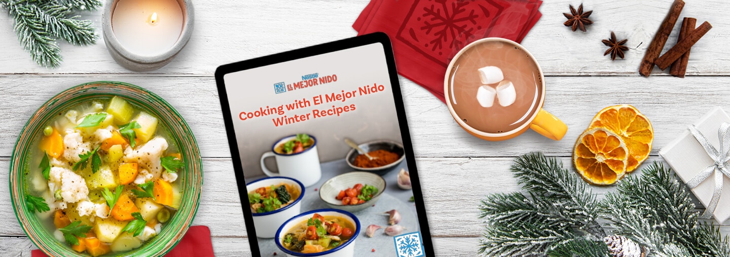 Tablet with Winter recipes next to hot chocolate, red napkins, spices, oranges, candles, soup and a wreath on a wood surface.
