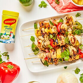 Sobrecarga de una botella amarilla Maggi sobre mesa blanca junto a un plato lleno de kebabs de gambas e ingredientes sueltos.