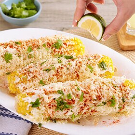 Primer plano de una mano exprimiendo una lima en tres mazorcas de maíz al estilo mexicano en un plato blanco.