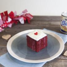 Slice of red velvet cake with white icing and a red decorative heart on top, on a light blue plate on a gray table.