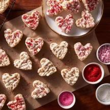Overhead of a dozen heart shaped cereal treats on a wood cutting board and ceramic plate all on a wood surface.